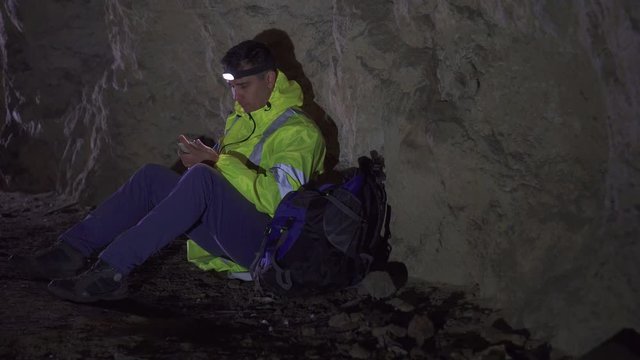Man tourist with a flashlight on his forehead and a phone in his hand lost in the cave, sitting and trying to call rescuers