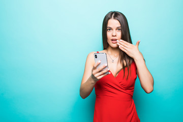 Portrait if a shocked young girl in dress looking at mobile phone isolated over blue background