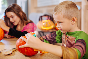 Friends and pumpkins. Blonde-haired cute boy wearing Halloween costume sitting near his friends coloring pumpkin