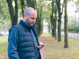 man is holding a phone, walking in the city Park, among the trees and looking at the smartphone