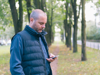 Fototapeta premium man is holding a phone, walking in the city Park, among the trees and looking at the smartphone
