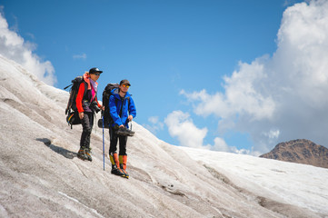 Fototapeta premium Two tourists, a man and a woman with backpacks and cats on their feet, stand on the ice in the background of the mountains of the sky and clouds. Watch in the distance