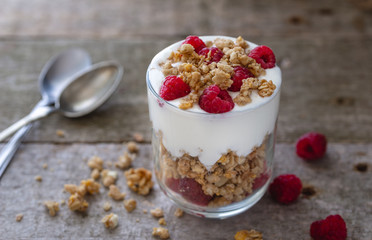 White yogurt with muesli in glass bowl with pieces raspberries on top on natural wooden desk.