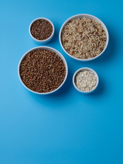 brown rice, buckwheat, flax and oat flakes in white cups on a blue background