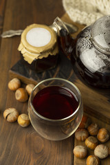 Tea in glass cup with teapot and knitted blanket near, with cinnamon sticks and hazelnut at wood background, with spoon and strainer near.