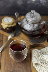 Tea in glass cup with teapot and knitted blanket near, with cinnamon sticks and hazelnut at wood background, with spoon and strainer near.