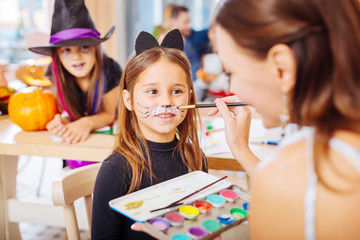 Obraz na płótnie Canvas Painting face. Dark-haired kindergarten teacher painting face for little dark-eyed girl wearing cat Halloween costume