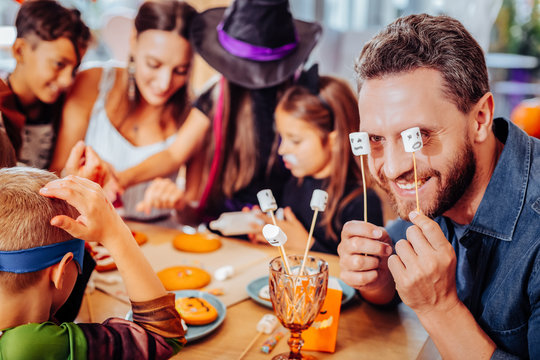 Marshmallows on sticks. Smiling bearded father holding marshmallows on sticks while celebrating Halloween with wife and children