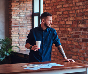 Handsome bearded man holds takeaway coffee and working with paper documents in the office with loft interior.