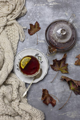 Cup with black tea and lemon and saucer, spoon, jelly in jar, maple leaves and knitted scarf near, an gray background