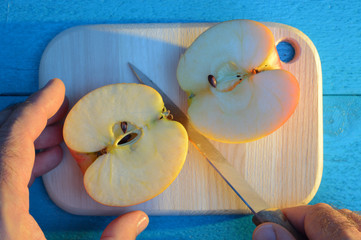 cutting an apple with his hand with knife on cutting wooden cutting board on a turquoise background at sunset