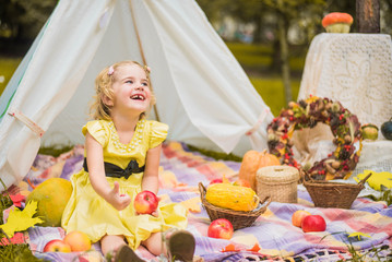 Little girl lying and playing in a tent, children's house wigwam in park Autumn portrait of cute curly girl. Harvest or Thanksgiving. autumn decor, party, picnic. Child in yellow dress with apple  © MartaKlos