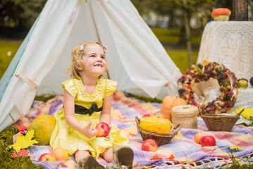 Little girl lying and playing in a tent, children's house wigwam in park Autumn portrait of cute curly girl. Harvest or Thanksgiving. autumn decor, party, picnic. Child in yellow dress with apple  © MartaKlos
