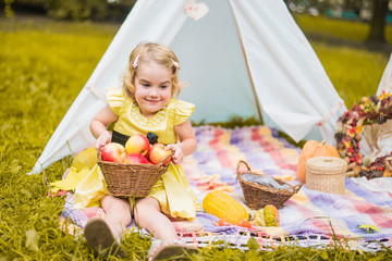 Little girl lying and playing in a tent, children's house wigwam in park Autumn portrait of cute curly girl. Harvest or Thanksgiving. autumn decor, party, picnic. Child in yellow dress with apple  © MartaKlos