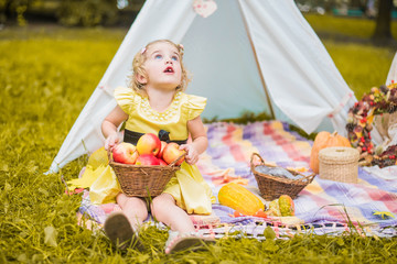 Little girl lying and playing in a tent, children's house wigwam in park Autumn portrait of cute curly girl. Harvest or Thanksgiving. autumn decor, party, picnic. Child in yellow dress with apple  © MartaKlos