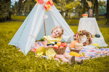 Little girl lying and playing in a tent, children's house wigwam in park Autumn portrait of cute curly girl. Harvest or Thanksgiving. autumn decor, party, picnic. Child in yellow dress with apple  © MartaKlos