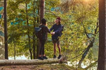 Fototapeta premium Young hipster couple holding hands standing on a tree trunk in a beautiful forest at sunset. Travel, tourism, and hike concept.