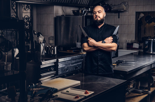 Bearded cook in black uniform holds knife and standing with crossed arms in the kitchen.