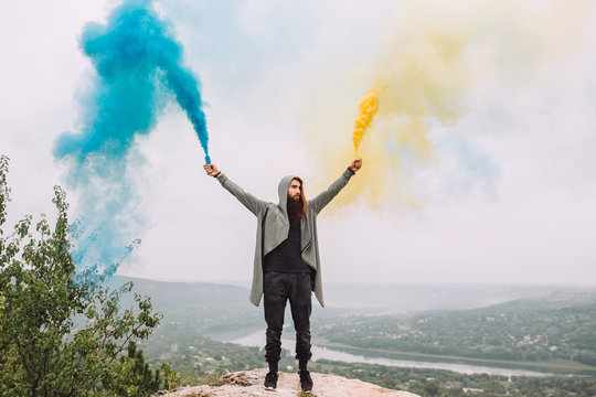 Fashionable Hipster Man Holding Colorful Smoke Bombs In Nature by