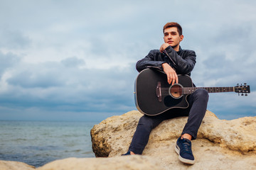 Young man with acoustic guitar sitting on beach surrounded with rocks on rainy day