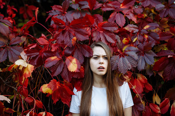 Bright autumn portrait. Young attractive blonde with long hair in a white t-shirt walks in the Park. She expresses her look of surprise and discontent against the background of colorful red foliage