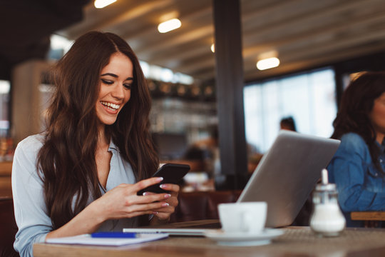 Young Woman Texting On The Phone With A Laptop On Table In Cafe