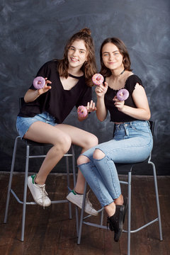 Pretty happy teenage girls with donuts  siting on chairs and having fun. Portrait of joyful smiling girls with donuts on black background. Good mood, diet concept.