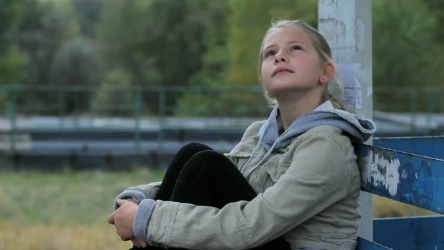 Cute preteen girl sitting alone hugging her knees daydreaming outdoors. Lonely young blonde teen girl thinking looking up.