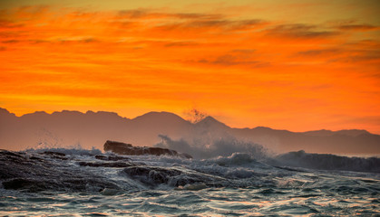 Seascape.  Clouds in a red dawn sky, waves crashing with splashes against stones, silhouettes of mountains on the horizon. False bay. South Africa.