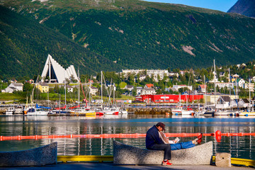 A couple sits on a bench in front of the calm, picturesque harbor at Tromso in far northern Norway in summer, with colorful boats and the Arctic Cathedral in the background