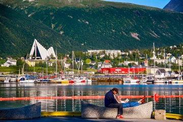 Fototapete Rund A couple sits on a bench in front of the calm, picturesque harbor at Tromso in far northern Norway in summer, with colorful boats and the Arctic Cathedral in the background © Kylie