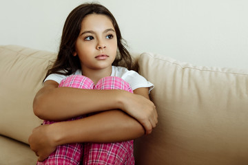 Loneliness. Home. Childhood. Sad girl is hugging her knees and looking away while sitting on couch at home