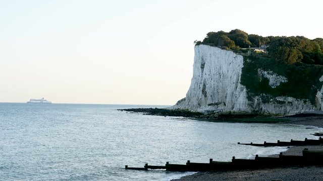Waves roll gently ashore with the iconic white cliffs of Dover in the evening shadows with a ferry boat in the far distant background.