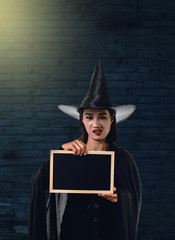 Woman in Black Scary witch halloween costume, Holding chalk board with Old Brick Wall of the Ancient Castle of the Witch