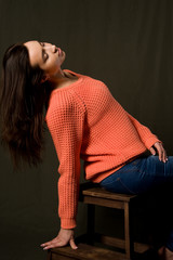 A beautiful young brunette girl in an orange knitted sweater poses against a dark background.
