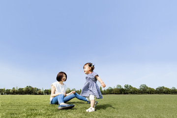 青空をバックに芝生の公園で幼い女の子と遊ぶお母さん。幸せ、育児、成長、健康、親子イメージ 