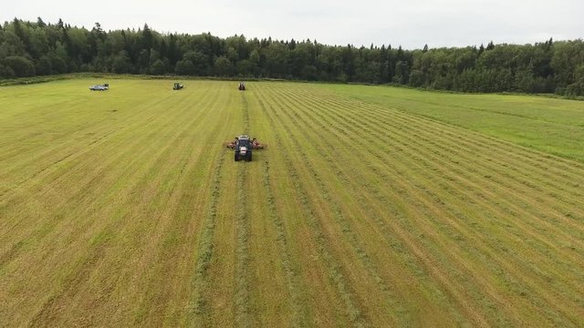 Drone camera is illustrating big grey tractor moving with separated attachment of red round shaped raker raking mowed hay on surface of large green farmland field.
