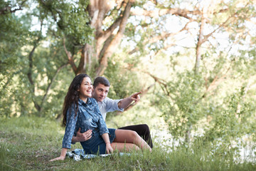 young couple sitting on the grass in the forest and looking on sunset, summer nature, bright sunlight, shadows and green leaves, romantic feelings