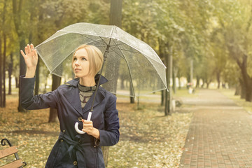 A woman of 30-40 years in a blue cloak goes under a transparent umbrella in the park.