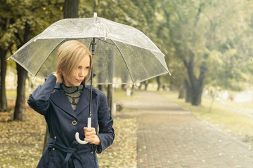 A woman of 30-40 years in a blue cloak goes under a transparent umbrella in the park.