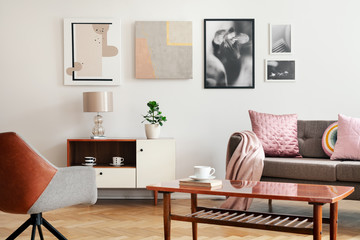 Real photo of white sitting room interior with poster on wall, couch with cushions and blanket, wooden coffee table with book and cup and plant on cupboard