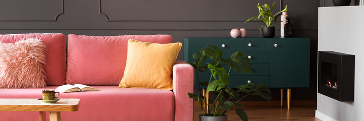 Panorama of orange pillow on pink settee in living room interior with plants and fireplace. Real...