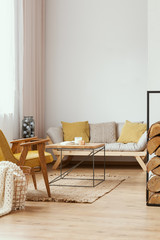 Pillows on couch and table on rug in white apartment interior with yellow wooden armchair. Real photo