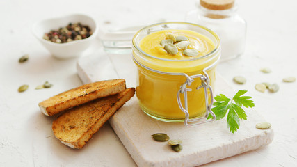 Yellow pumpkin soup served in glass jar with bread placed on chopping board on white background
