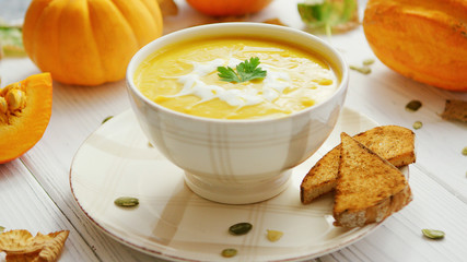 Yellow creamy pumpkin soup in bowl with pieces of bread surrounded by pumpkins on wooden table