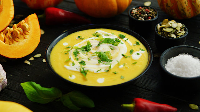 From Above View Of Yellow Pumpkin Soup In Black Bowl Sprinkled With Green Herbs Surrounded With Vegetables And Spices On Wooden Table