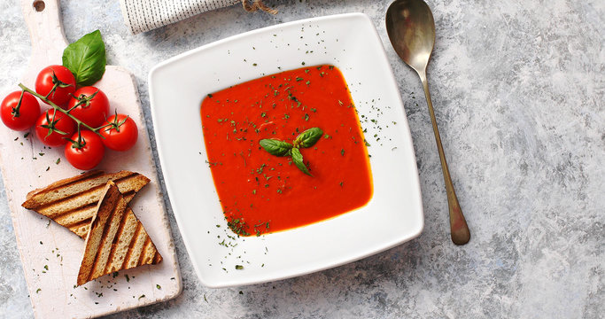 From above view of red tomato soup in white plate served with crisp bread and tablespoon on gray background 