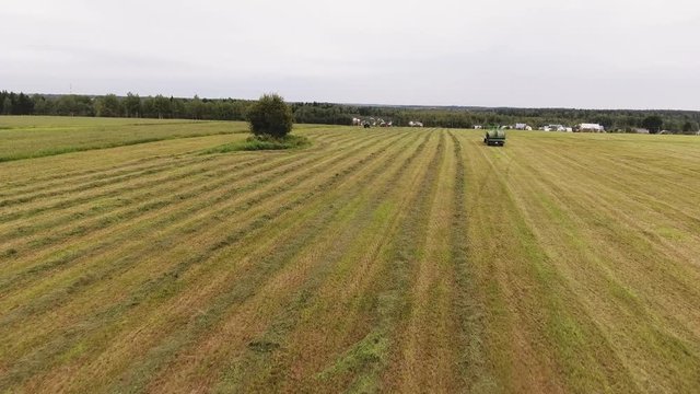Drone camera is showing green detached long trailer with round shaped hay bales on large yellow plowed farmland field surrounded by trees.