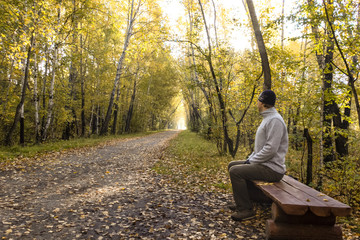 Fototapeta premium A lonely, warmly dressed man sits in an autumn park and looks thoughtfully at the road, thinking about something, or someone waiting.