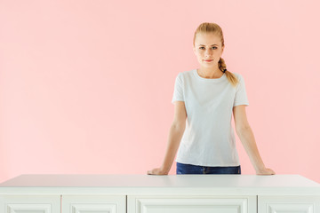attractive young woman standing behind kitchen table isolated on pink
